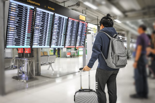 Asian Man Waiting Flight With A Luggage Looking At Departures Board In Airport
