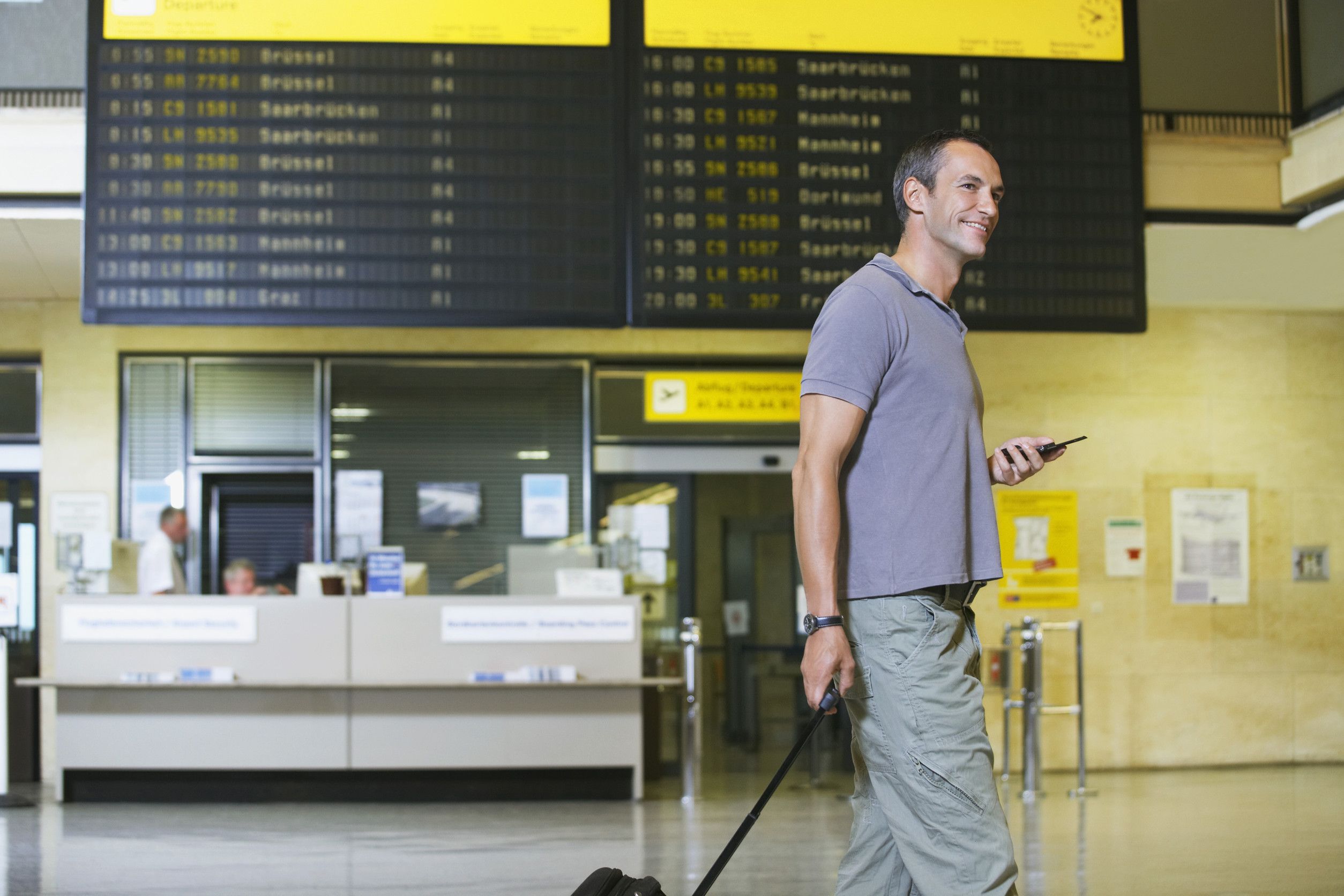 Traveller Holding Mobile Phone In Front Of Flight Status Board In Airport